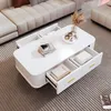 A white coffee table with drawers is placed in a living room setting, next to a sectional sofa. Produktbild-Vorschau