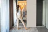 A woman is standing in front of an open wardrobe, looking at the clothes inside. Podgląd zdjęcia produktu