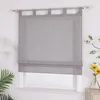 A gray Roman blind hangs on a window, with a lamp and plants visible to the side. Produktbild-Vorschau