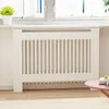 A white radiator cover with vertical slats is shown in a room with a window and a pink table in the foreground. Podgląd zdjęcia produktu
