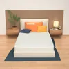 A mattress is displayed on a bed frame, with pillows and a bedside table with a lamp and decorative items. Produktbild-Vorschau