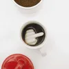 A white mug filled with dark coffee, featuring a white ceramic middle finger figurine submerged in the coffee. A white bowl filled with instant coffee granules is positioned above the mug, and a red lid is placed to the bottom left of the mug, all on a whi Produktbild-Vorschau