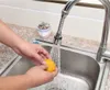 A person is washing an orange under a flexible faucet extender in a kitchen sink. Produktbild-Vorschau