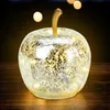 A decorative glass apple with LED lights sits on a wooden surface, with blurred lights in the background. Voir les images du produit