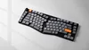 A split ergonomic keyboard with gray and orange keys is shown on a white and light gray surface with shadows. Produktbild-Vorschau
