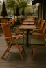 An outdoor seating area with wooden chairs and tables, some with green cushions. Produktbild-Vorschau