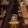 A close-up view of a miniature book nook with a small framed picture on a stool, set against a backdrop of shelves filled with books and decorative items. Náhled obrázku produktu