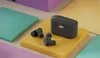 A pair of gray wireless earbuds and their charging case are displayed on a yellow platform, surrounded by colorful textured blocks. Produktbild-Vorschau