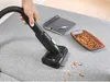 A hand is using a black Bosch vacuum cleaner to clean up crumbs on a gray fabric surface, next to a notebook and a bowl of pretzels. Produktbild-Vorschau