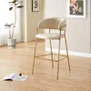 A beige bar stool with gold legs is placed in a room with light wood flooring, a potted plant, and striped wallpaper. Produktbild-Vorschau