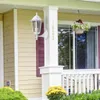 A close-up view of a white outdoor wall lamp mounted on a white pillar, with yellow flowers in the foreground and a window with blinds in the background. Produktbild-Vorschau