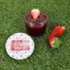 A jar of strawberry jam with a strawberry on top, and two whole strawberries next to it, all placed on artificial grass. A lid with a heart design and text is also visible. Náhled obrázku produktu