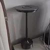 A dark gray LED table lamp with a circular base and top, standing on a white surface next to a chair. Produktbild-Vorschau
