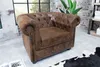 A brown Chesterfield armchair is shown in a room with a brick wall and a large window. Náhled obrázku produktu