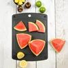 A black cutting board with slices of watermelon, limes, mangosteens, and lemons on a white wooden background. Produktbild-Vorschau