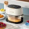 A cream-colored air fryer with a transparent glass bowl sits on a kitchen counter, surrounded by fresh produce and kitchen utensils. Produktbild-Vorschau