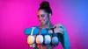 A woman is posing with multiple headphones around her arm, against a vibrant pink and blue gradient background. Produktbild-Vorschau