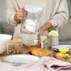 A person is holding a water filter pitcher over a table set for breakfast with croissants and a glass of water. Produktbild-Vorschau