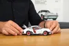 A person's hands are shown holding a white model car on a wooden table. A poster of a similar car is visible in the background. Náhled obrázku produktu