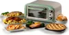 A retro-style mini oven is displayed with various prepared dishes surrounding it, including a roast, chicken, salmon, bread rolls, and a pie. Anteprima dell’immagine prodotto