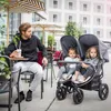 A man sits at a table with a drink while a double stroller with two children is next to him. Produktbild-Vorschau