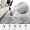 A fluffy gray rug is laid out on a wooden floor in a living room, with a white sofa and a potted plant in the background. A laptop is placed on the rug. Náhled obrázku produktu