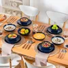 A dining table set with blue ceramic dishes filled with various foods, including soup, cereal, and fruit, with chairs in the background. Náhled obrázku produktu