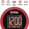 A close-up of a red alarm clock with a digital display showing the time 12:00 PM. Above the display is the brand name 'Wake'n Shake DYNAMITE'. Along the top of the image are icons representing features like alarm, night light, USB charging, brightness, and Voir les images du produit