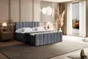 A bedroom scene featuring a dark gray velvet boxspring bed with a headboard, two pillows, and a mattress with a topper. A bedside table with a lamp is to the left, and a large window with curtains is to the right. Decorative dried plants and a wall light a Produktbild-Vorschau