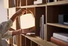 A person's hands are holding a pink and white portable lamp with a handle, placing it on a wooden shelf next to books. Produktbild-Vorschau