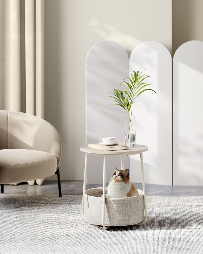 A cat is sitting in a round pet bed integrated into a side table, with a vase of greenery and a teacup on top. The scene is set in a modern, minimalist living room with soft lighting and neutral tones.
