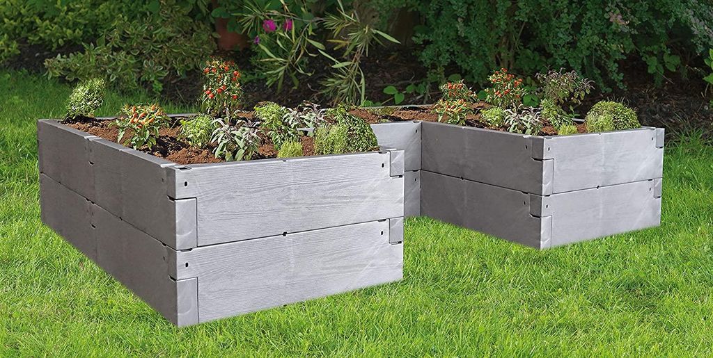 Two raised garden beds filled with plants, set on a grassy lawn with a garden in the background.