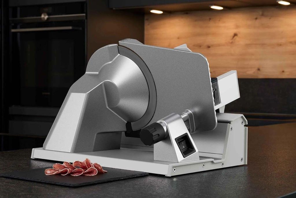 A silver meat slicer is shown on a dark countertop, with sliced meat in front of it. The background shows kitchen cabinets and appliances.