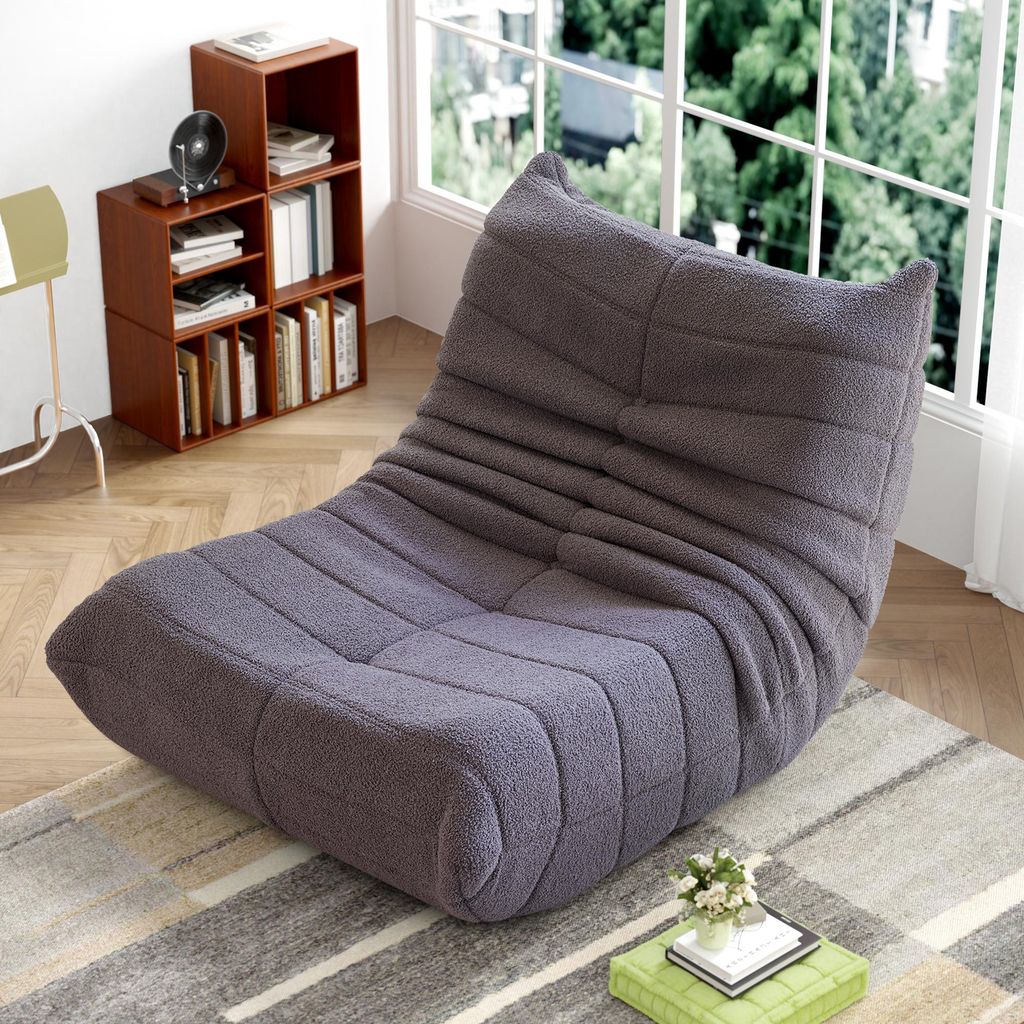 A gray floor chair with a textured fabric is placed on a rug in a room with a bookshelf and a window.