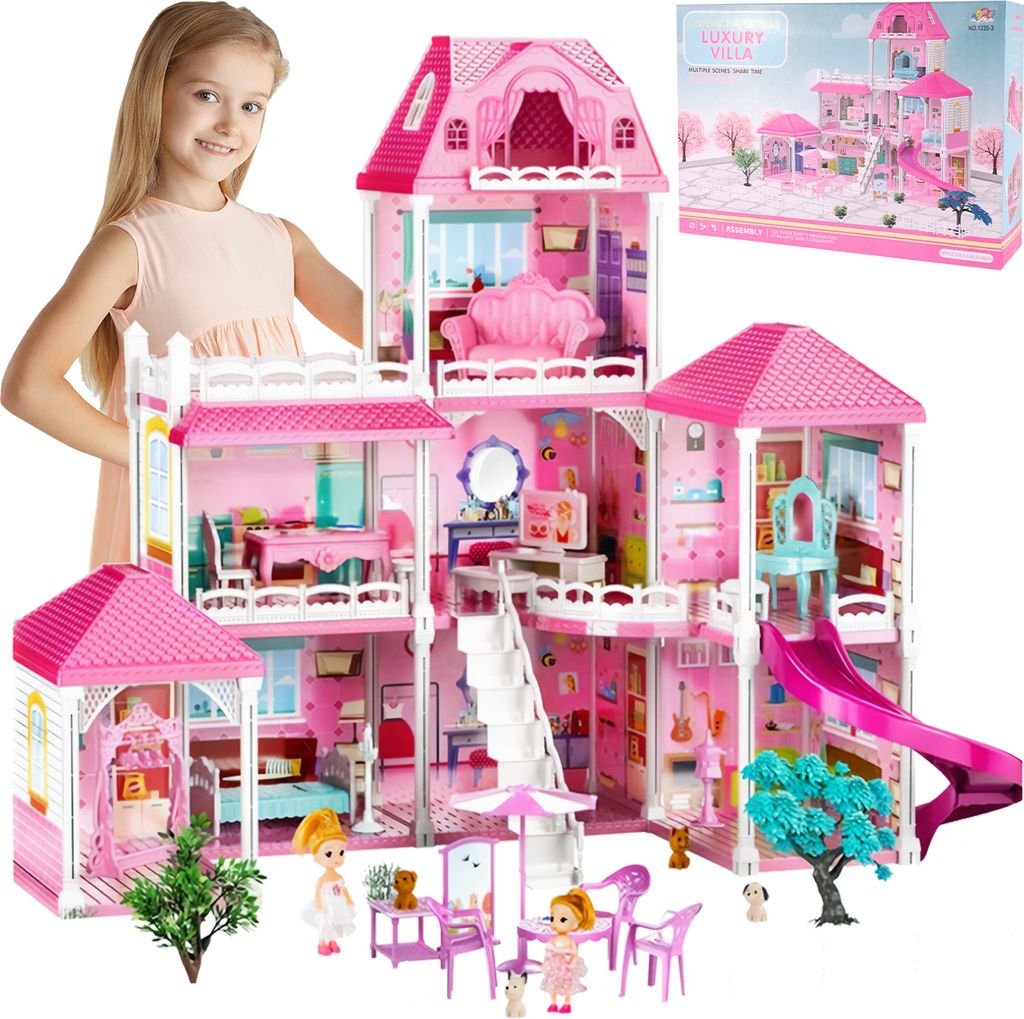 A young girl stands behind a large pink dollhouse, which is set up with furniture, dolls, and accessories. The dollhouse is elaborate and multi-story, featuring a slide, balconies, and various rooms. A box for the dollhouse is visible in the upper right co