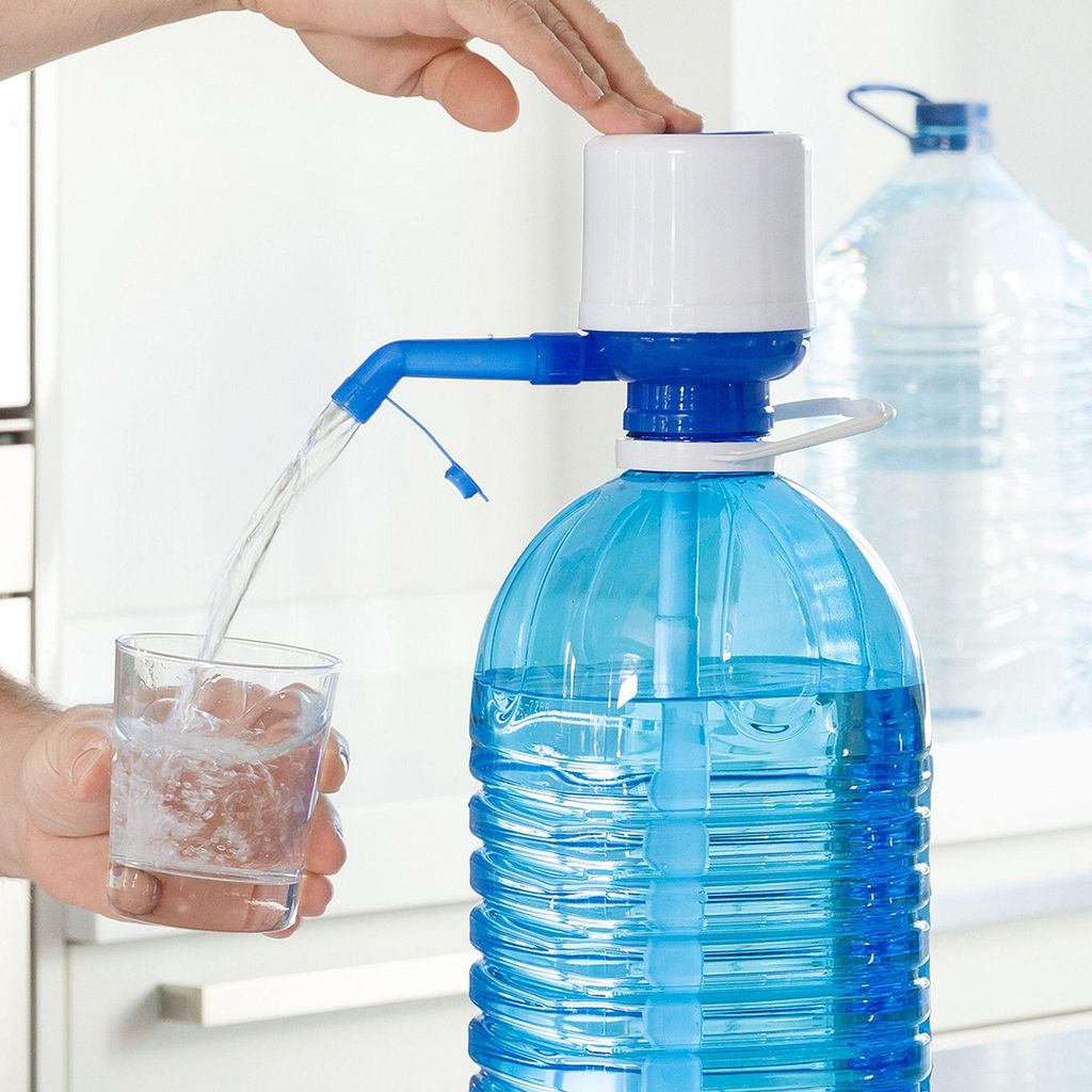 A hand is pressing down on a water dispenser pump attached to a large blue water bottle, dispensing water into a small glass. Another large water bottle is visible in the background.
