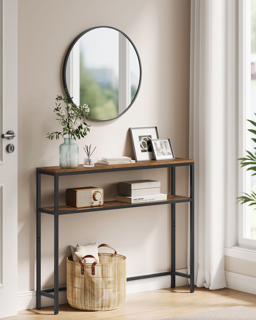 A narrow console table with two shelves is placed in a hallway, decorated with a mirror, framed pictures, a radio, books, and a woven basket.