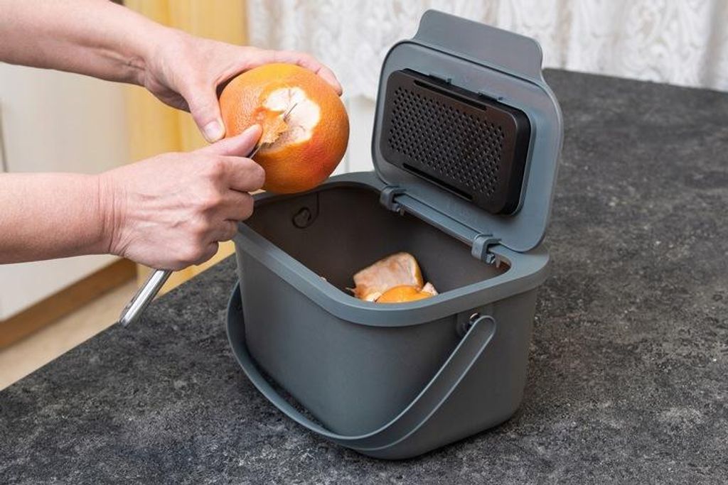 A person is peeling an orange over a compost bin.