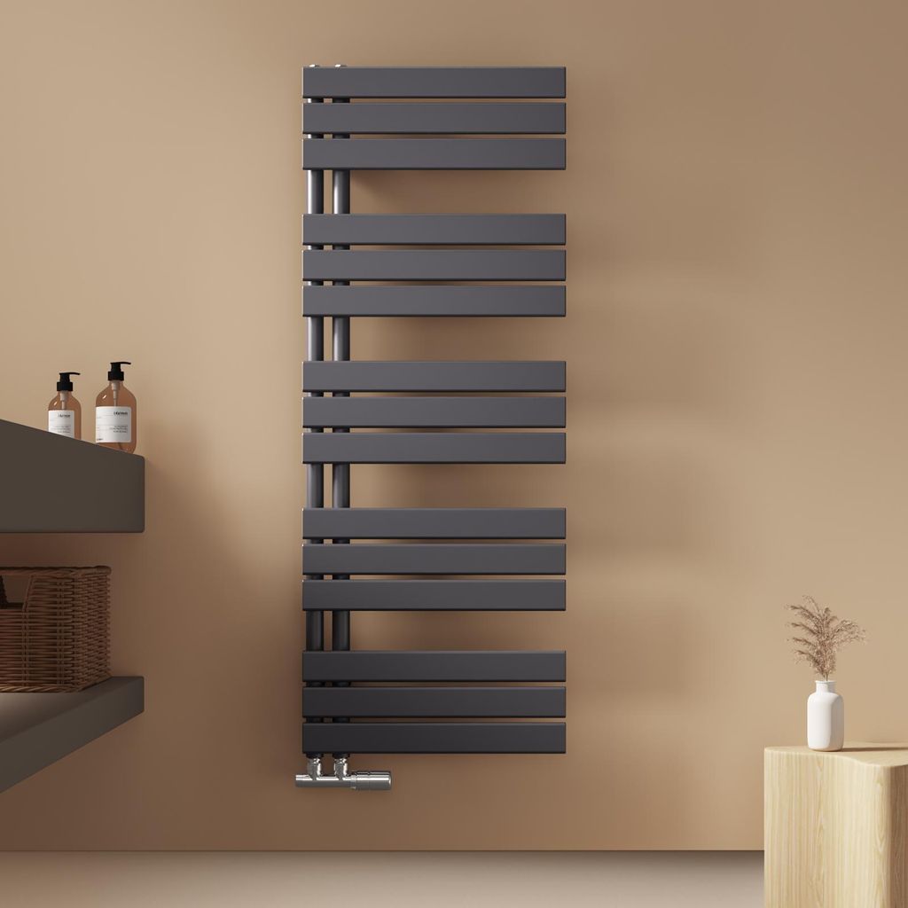 A modern anthracite towel radiator mounted on a light brown wall, with a shelf and soap dispensers to the left, and a small decorative plant on a wooden cube to the right.