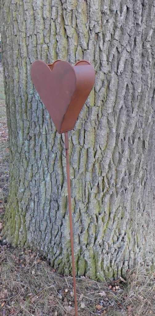 A rusty metal heart garden stake is placed next to a tree trunk.