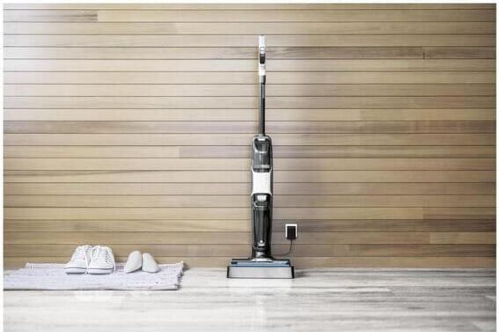 A vacuum cleaner is standing upright against a wood-paneled wall, with a pair of shoes on a mat in front of it.