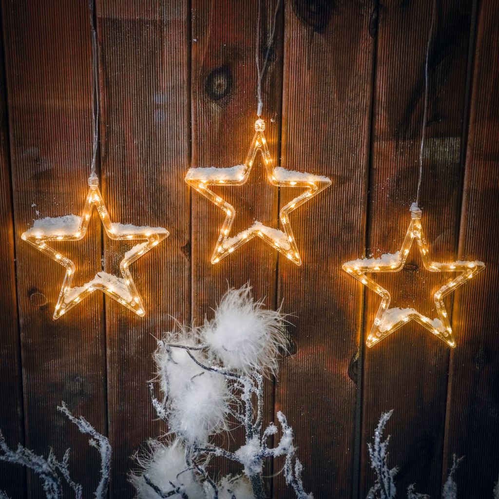 Three illuminated star-shaped lights with artificial snow hang against a rustic wooden wall, with frosted branches and fluffy white decorations in the foreground.