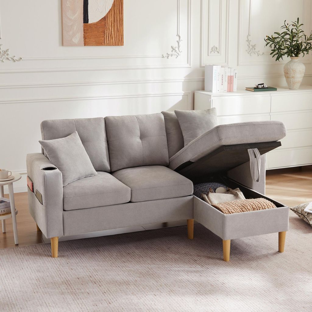 A light gray sectional sofa with its storage ottoman open, revealing stored items. The sofa is in a well-lit living room with decorative elements on the wall and a dresser in the background.