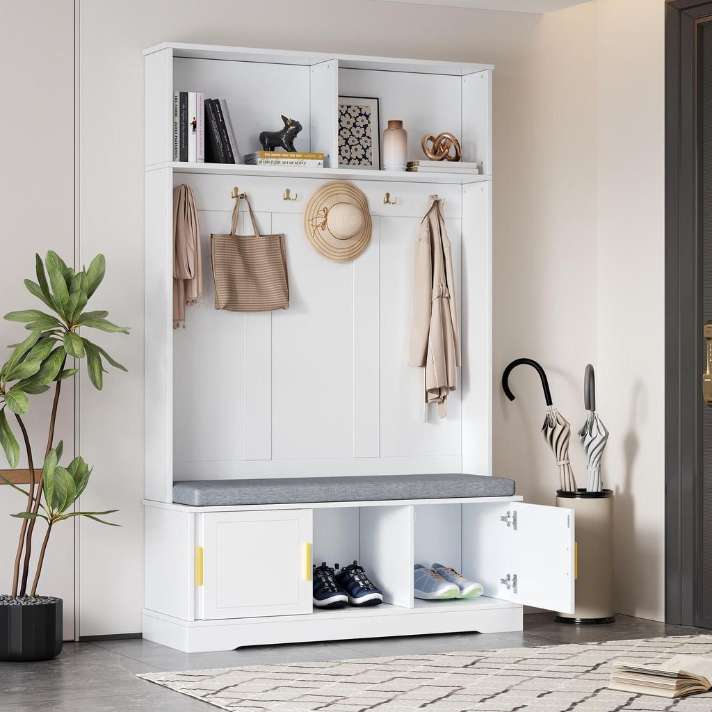 A white entryway hall tree with a bench and storage compartments, featuring hooks for coats and bags, and shelves for shoes. It is placed in a home entryway with a potted plant and an umbrella stand nearby.