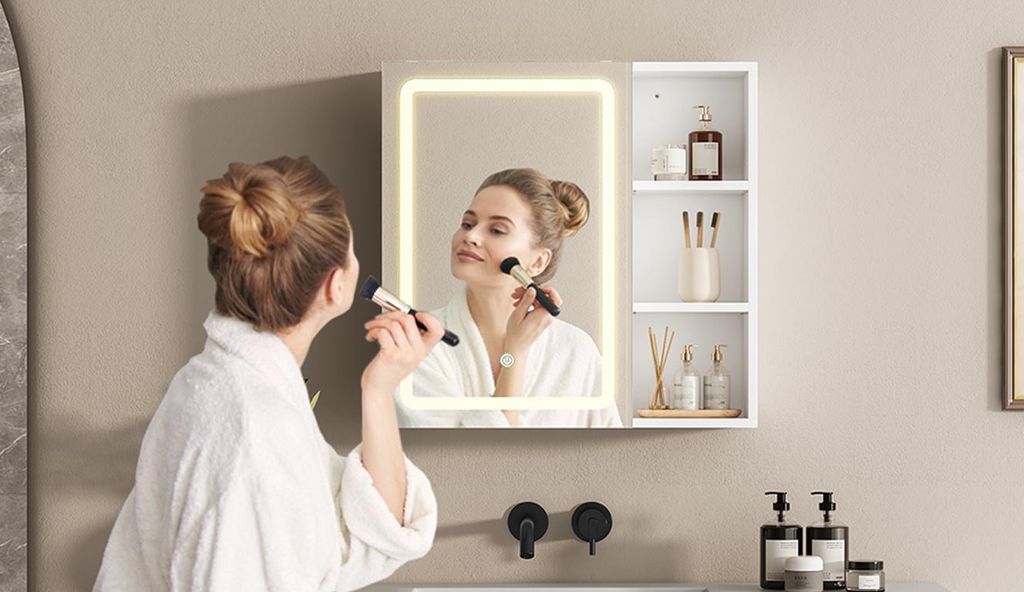 A woman applying makeup in front of a lighted mirror cabinet with shelves.
