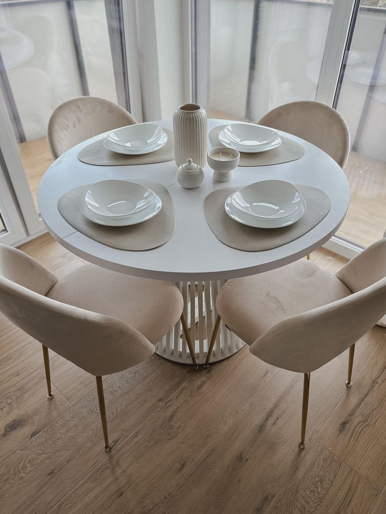 A round white dining table with four beige chairs and placemats with bowls set for a meal.