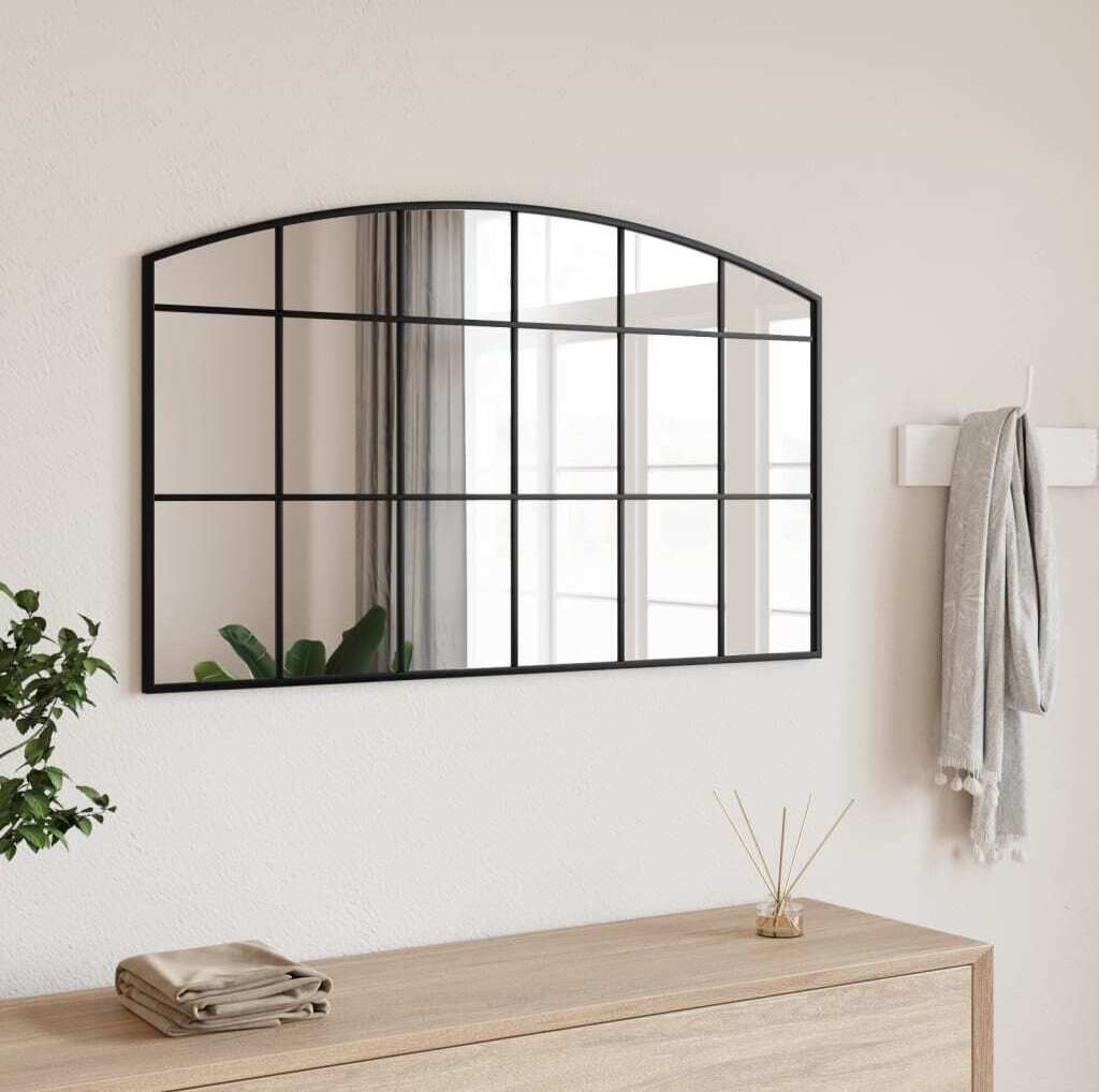 A black arched mirror with a grid design hangs on a white wall above a wooden dresser. A gray scarf is draped over a hook to the right of the mirror, and a small plant and diffuser are on the dresser.