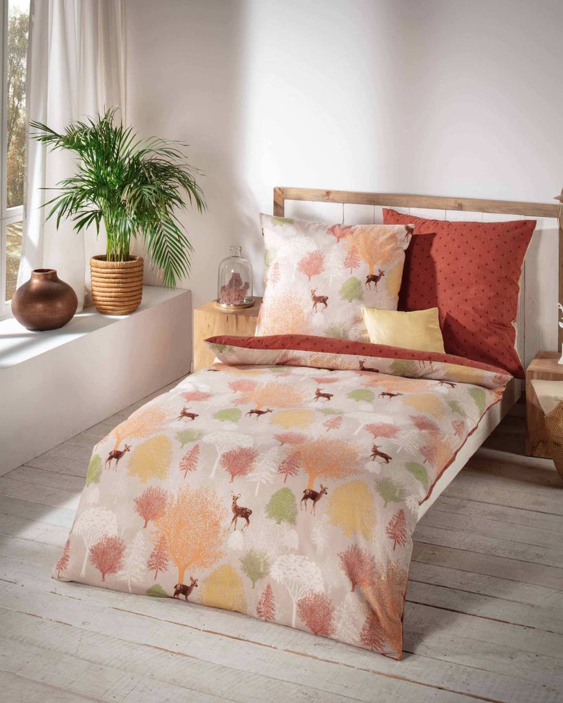 A bed with bedding featuring a forest and deer pattern, set in a room with a window and plants.