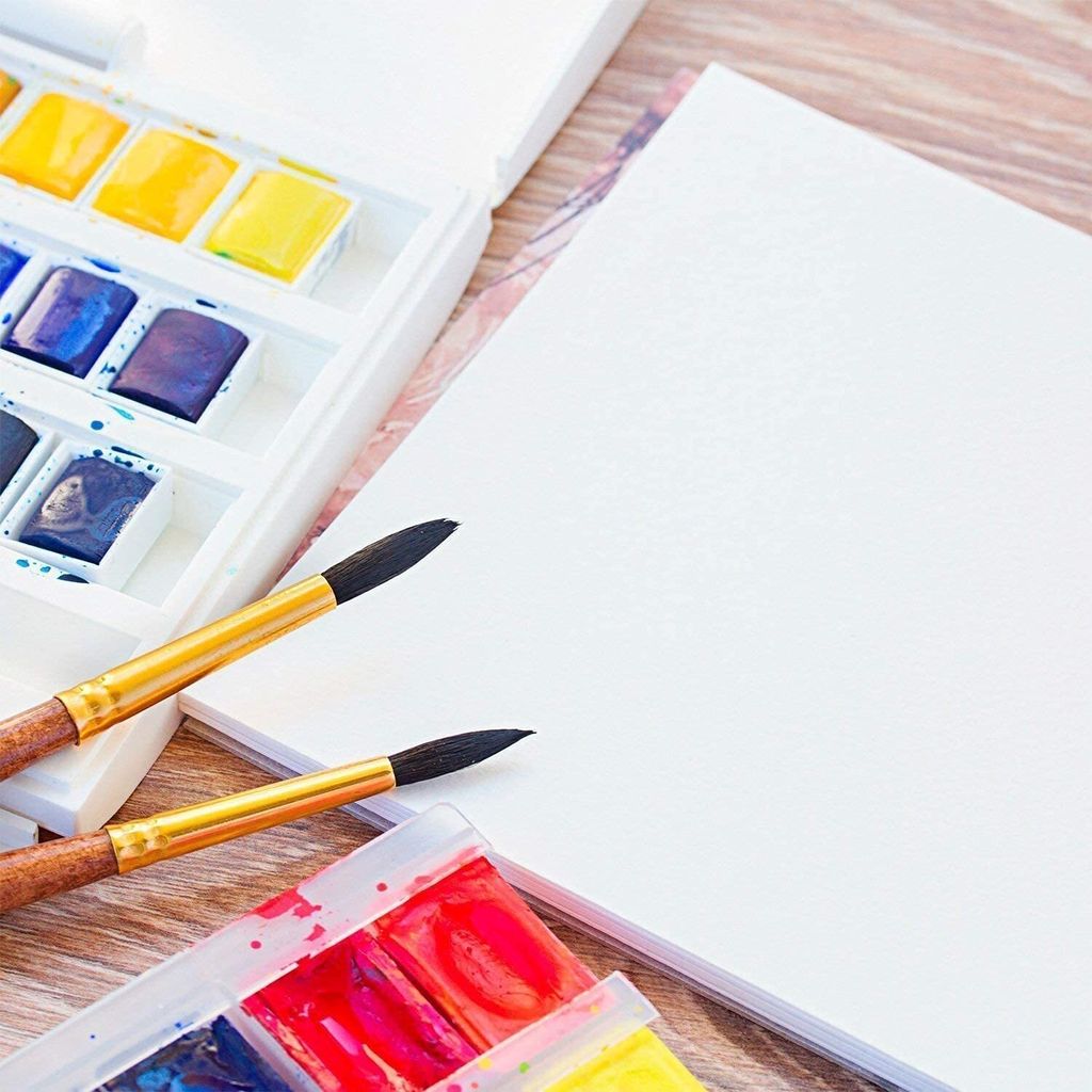 A close-up overhead view of art supplies including a watercolor paint set, brushes, and a blank sheet of paper on a wooden surface.