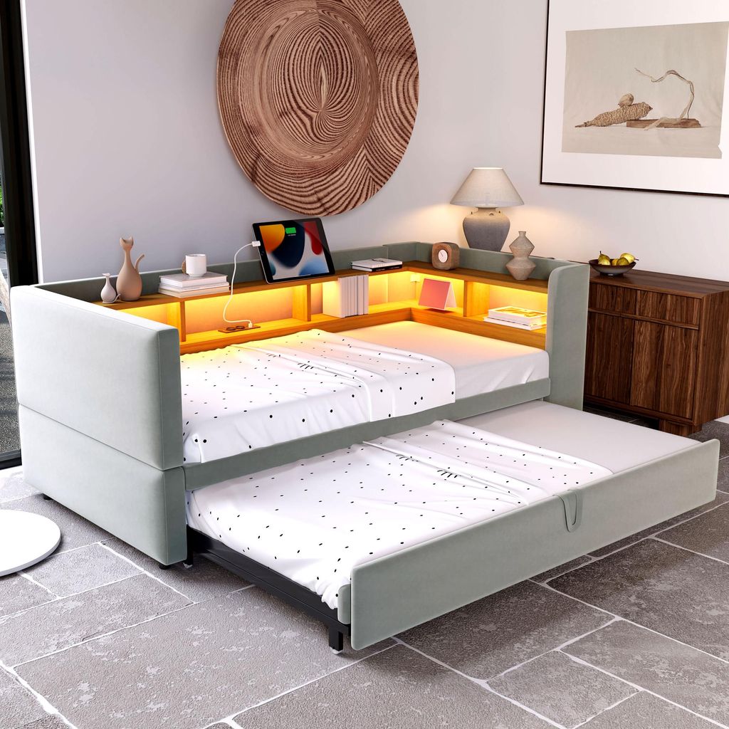 A modern living room or bedroom with a light gray sofa bed extended with a pull-out mattress, featuring built-in LED lighting and shelves with decorative items and a tablet.